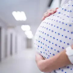 Kobieta w szpitalnej koszuli z opaską na ręku obejmuje brzuch. W tle szpitalny korytarz, może symbolizować stres w ciąży.