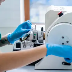 Ręce w niebieskich rękawiczkach pracują przy mikrotomie, przygotowując próbkę do badania. To kluczowy etap w procesie diagnostycznym, jakim jest biopsja.