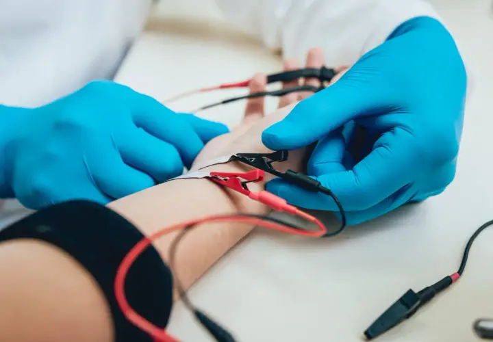 Lekarz w niebieskich rękawiczkach zakłada elektrody na rękę pacjenta. To badanie EMG, które pozwala ocenić funkcję nerwów i mięśni.