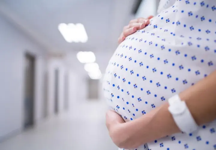 Kobieta w szpitalnej koszuli z opaską na ręku obejmuje brzuch. W tle szpitalny korytarz, może symbolizować stres w ciąży.