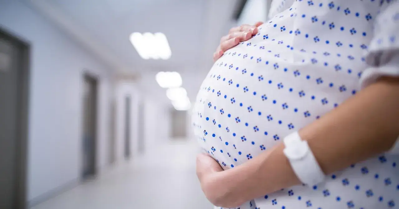 Kobieta w szpitalnej koszuli z opaską na ręku obejmuje brzuch. W tle szpitalny korytarz, może symbolizować stres w ciąży.