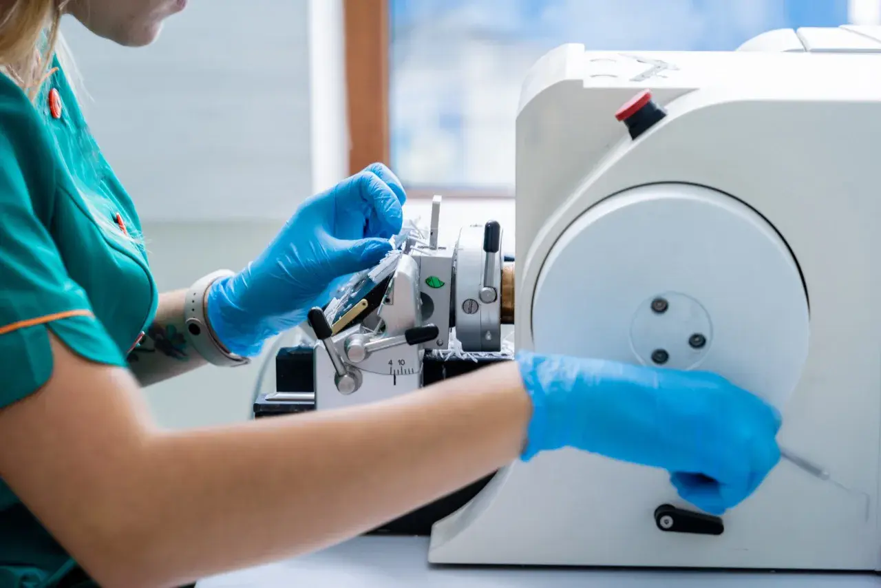 Ręce w niebieskich rękawiczkach pracują przy mikrotomie, przygotowując próbkę do badania. To kluczowy etap w procesie diagnostycznym, jakim jest biopsja.
