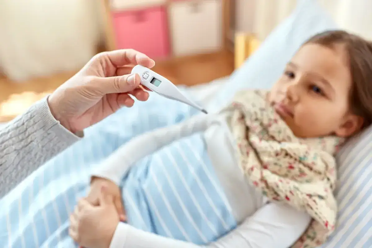 Dziecko leży w łóżku z gorączką. Ręka trzyma termometr, a obok leży paracetamol dla dzieci.